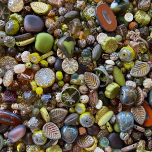 May include: A large collection of assorted beads in shades of olive green, brown, yellow, and amber. The mix includes various shapes such as leaves, flowers, round spheres, and flat discs, with some beads featuring dragonfly and floral patterns.