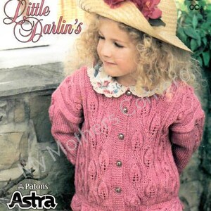 Könnte beinhalten: Ein rosa gestrickter Cardigan mit einem Zopfmuster und einem Knopfverschluss. Der Cardigan hat einen gewellten Saum und wird von einem jungen Mädchen mit lockigem Haar getragen, das einen Strohhut mit Blumen trägt.