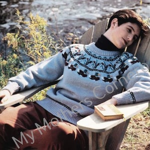 May include: A light blue jumper with a colourful geometric pattern around the neck. The jumper is being worn by a person sitting in a wooden chair.