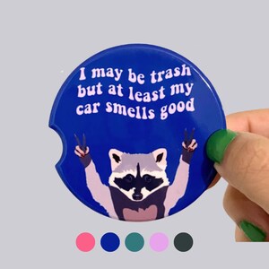 May include: A circular blue car coaster featuring a graphic of a raccoon making peace signs with its paws. The text printed in white reads, I may be trash but at least my car smells good. The coaster has a small semi-circular notch on the left side.