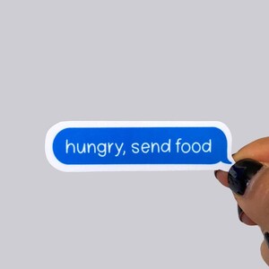 May include: A blue and white sticker in the shape of a text message bubble. The text "hungry, send food" is written in white, sans-serif font. The sticker is held by a hand with black nail polish.