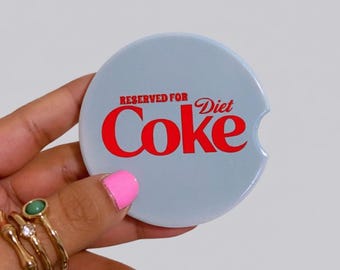 以下が含まれることがあります： 前面に赤い文字で「RESERVED FOR Diet Coke」とプリントされた、円形の水色の車用カップホルダーコースター。コースターの右側には、車用カップホルダーから取り出しやすいように小さな半円形の切り込みが入っています。