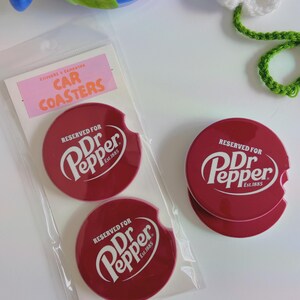 May include: Three red car coasters featuring the Dr Pepper logo in white. The coasters are circular with a convenient notch. The packaging includes a pink label that reads "STICKERS & SAMASTRA CAR COASTERS".