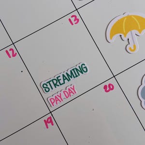 May include: A white calendar page with pink numbers 12, 13, 14, 19, and 20. A sticker with the green text STREAMING and pink text PAY DAY is placed on the 19th. Other stickers include a yellow umbrella and a blue cloud on the calendar grid.
