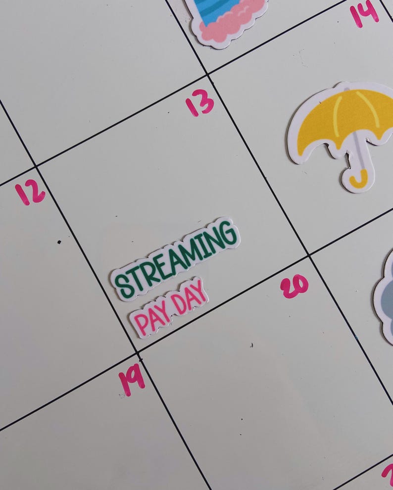 May include: A white calendar page with pink numbers 12, 13, 14, 19, and 20. A sticker with the green text STREAMING and pink text PAY DAY is placed on the 19th. Other stickers include a yellow umbrella and a blue cloud on the calendar grid.