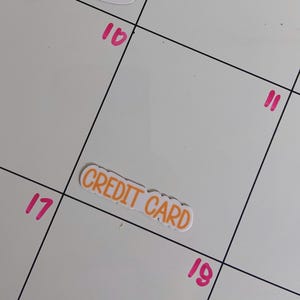 May include: A white rectangular sticker with the words CREDIT CARD printed in orange capital letters. The sticker is placed on a white calendar grid featuring black lines and pink numbers 10, 11, 17, and 19, with a partial pink and tan sticker above.