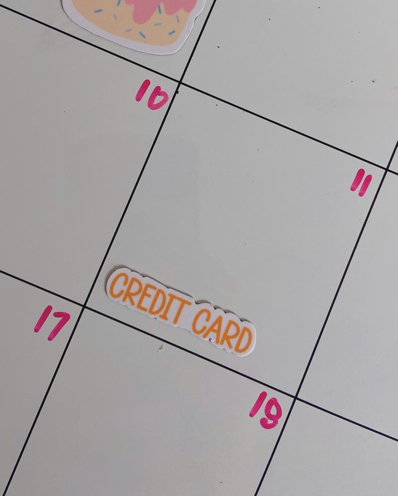 May include: A white rectangular sticker with the words CREDIT CARD printed in orange capital letters. The sticker is placed on a white calendar grid featuring black lines and pink numbers 10, 11, 17, and 19, with a partial pink and tan sticker above.