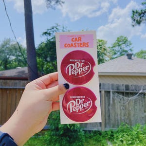 May include: Two burgundy car coasters with the Dr. Pepper logo in white. The coasters are round and are presented on a pink and white card, which reads "Car Coasters".
