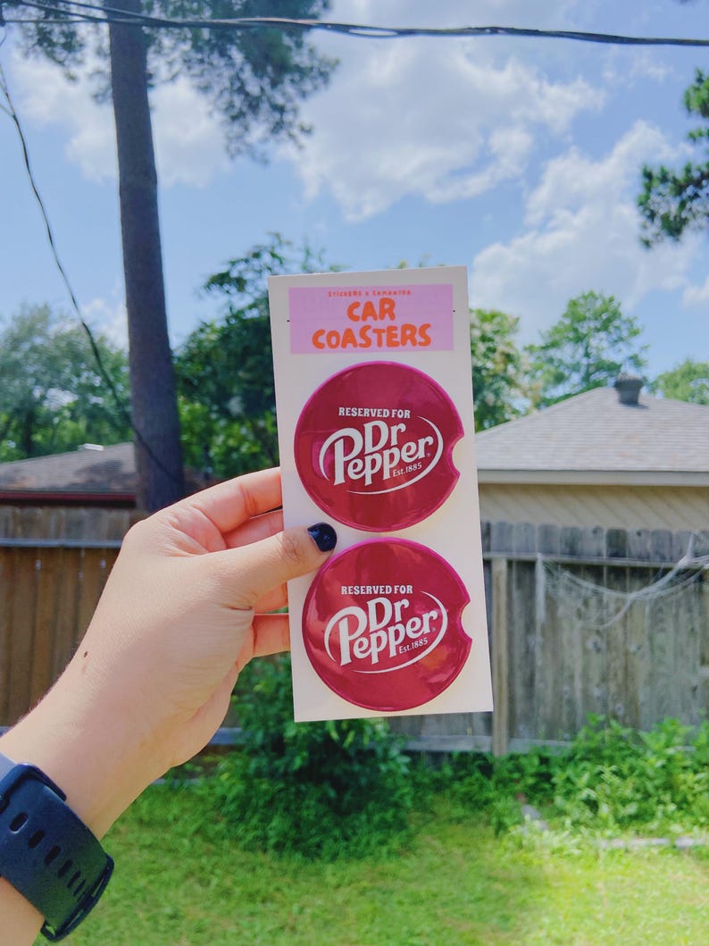 May include: Two burgundy car coasters with the Dr. Pepper logo in white. The coasters are round and are presented on a pink and white card, which reads "Car Coasters".