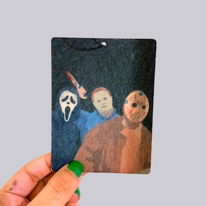 May include: A rectangular air freshener featuring a horror movie theme. The design includes illustrations of characters from the films Scream, Halloween, and Friday the 13th. The background is dark, and the characters are in color.