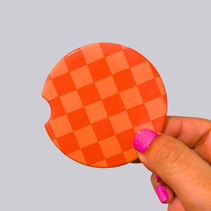 May include: A circular car coaster featuring a vibrant orange and light orange checkered pattern. The coaster has a small, semi-circular notch cut out from the left side of its perimeter, designed for easy removal from a vehicle cup holder.