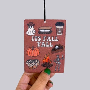 May include: A rectangular air freshener with a brown background and black string. The air freshener features fall-themed illustrations including a ghost, s'mores, coffee, pumpkin, pie, cinnamon rolls, a campfire, and a skeleton. The text "ITS FALL YALL" is in the center.
