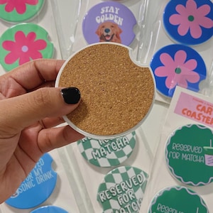 May include: Car coasters with diverse designs, including a flower, a golden retriever with "Stay Golden", and "Reserved for Matcha". The coasters are packaged in clear plastic. The coasters are designed to fit in a car's cup holder.