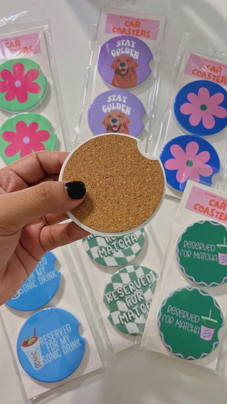May include: Car coasters with diverse designs, including a flower, a golden retriever with "Stay Golden", and "Reserved for Matcha". The coasters are packaged in clear plastic. The coasters are designed to fit in a car's cup holder.