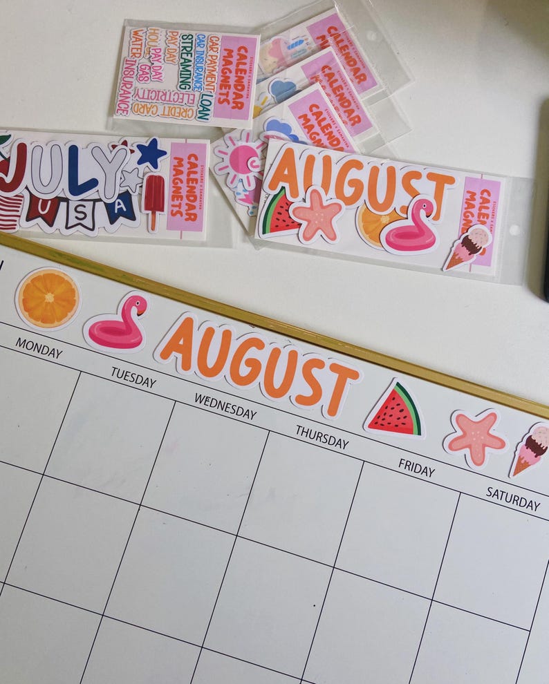 May include: A white magnetic calendar board with a gold frame, featuring the word AUGUST in orange letters and summer-themed magnets including a flamingo, watermelon, and ice cream. Additional packs of calendar magnets are visible nearby.
