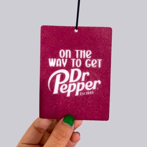 May include: A burgundy air freshener with the Dr. Pepper logo and the text "ON THE WAY TO GET Dr Pepper Est. 1885" in white. The air freshener is rectangular with a textured surface and a black string for hanging.