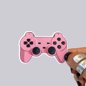 May include: A pink sticker of a video game controller. The controller has black buttons and directional pad. The sticker is held by a hand with silver rings and black nail polish.