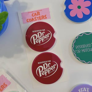 May include: Two red car coasters featuring the Dr. Pepper logo and the text "RESERVED FOR Dr Pepper Est. 1885". The coasters are round with a curved cutout. Other coasters with floral and text designs are also visible.