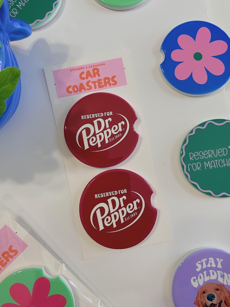 May include: Two red car coasters featuring the Dr. Pepper logo and the text "RESERVED FOR Dr Pepper Est. 1885". The coasters are round with a curved cutout. Other coasters with floral and text designs are also visible.