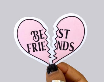 以下が含まれることがあります： 「BEST FRIENDS」の黒い文字が入った、割れたハート型のピンク色のステッカー。 ハートのモチーフが付いた2つのシルバーリングをつけた手で持っています。 背景は無地のライトグレーです。