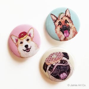 May include: Three round button pins featuring different dog breeds wearing food on their heads. The first pin features a corgi wearing a pink donut, the second pin features a German Shepherd wearing a green taco, and the third pin features a pug wearing a waffle.