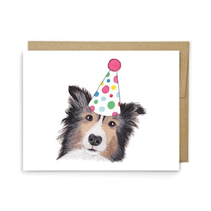 May include: A white greeting card featuring a watercolor illustration of a black, white, and tan dog wearing a conical party hat decorated with colorful polka dots. The card is paired with a plain brown envelope positioned directly behind it.