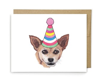 May include: A white greeting card featuring a watercolor illustration of a brown dog wearing a colorful striped party hat with a pink pom-pom on top. The dog has pointed ears, dark eyes, and a black nose, centered on the front of the card.