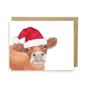 Puede incluir: Una tarjeta de felicitaci&oacute;n con una ilustraci&oacute;n en acuarela de una vaca marr&oacute;n que lleva un gorro de Pap&aacute; Noel rojo y blanco. La vaca tiene ojos grandes y oscuros y un hocico claro. La tarjeta est&aacute; sobre un fondo blanco con un sobre marr&oacute;n.