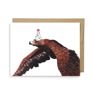 May include: A greeting card featuring a watercolor illustration of a brown eagle in flight, wearing a small, light blue party hat decorated with colorful polka dots. The card is positioned against a plain, light brown envelope background.