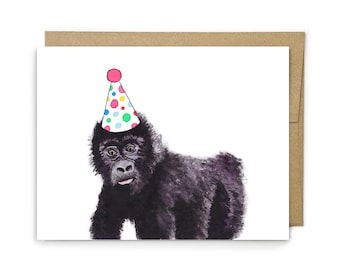 May include: A greeting card featuring a watercolor illustration of a black gorilla wearing a white party hat decorated with colorful polka dots. The card is positioned in front of a brown kraft paper envelope, creating a simple, clean visual.