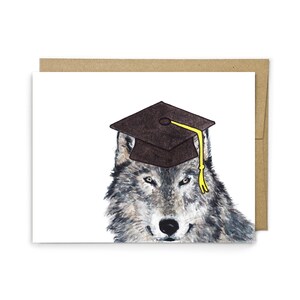May include: A greeting card featuring a watercolor illustration of a gray wolf wearing a dark brown graduation cap with a bright yellow tassel. The wolf has detailed fur textures in shades of gray, white, and brown, set against a plain white background.