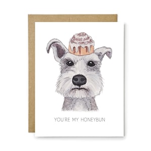May include: A white greeting card featuring a watercolor illustration of a gray and white schnauzer dog with a cinnamon roll balanced on its head. Below the dog, the text reads YOU'RE MY HONEYBUN in simple black capital letters.