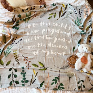 Può includere: Una coperta per bambini grigio chiaro con un design botanico con foglie e steli verdi, marroni e arancioni. La coperta ha il testo "For this child I have prayed and the Lord has granted me the desires of my heart." Un orsacchiotto e dei guanti sono sulla coperta.