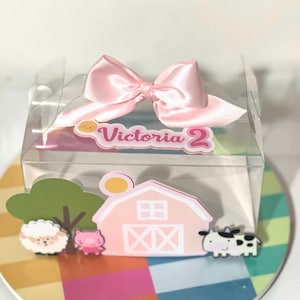 Puede incluir: Una caja de plástico transparente con un lazo rosa en la parte superior. La caja está decorada con una escena de granja, que incluye un granero, una vaca, un cerdo y una oveja. La caja también tiene una etiqueta que dice "Victoria 2".
