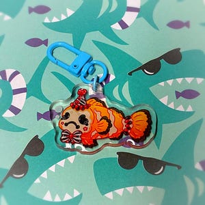 May include: A clear acrylic keychain featuring a cartoon clownfish with orange and white stripes, a party hat, and a bow tie. The keychain has a blue clasp. The background is a teal pattern with sharks, sunglasses, and fish.