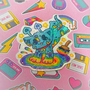 May include: A holographic sticker featuring a blue cartoon cat with rainbow paint dripping from its body, standing in a rainbow puddle. Surrounding the cat are other stickers with 90s-themed designs, including a cassette tape, floppy disks, and a Rubik's cube.