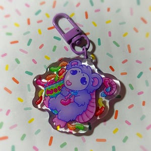 Puede incluir: Llavero de ratón de dibujos animados morado con cola rosa que sostiene una caja de gusanos gomosos. El llavero está hecho de acrílico transparente y tiene un cierre de metal morado.