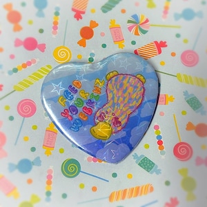 Puede incluir: Un botón azul en forma de corazón con un ornitorrinco de dibujos animados que lleva un atuendo estampado en rosa y amarillo. El ornitorrinco está rodeado de nubes blancas y estrellas. El texto "Feed Your Webkinz" está impreso en el botón.