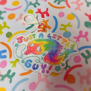 May include: A clear acrylic charm with a rainbow-colored creature design and the text "JUST A LITTLE GUY" in colorful letters. The charm has a white border and a lobster clasp for attachment.
