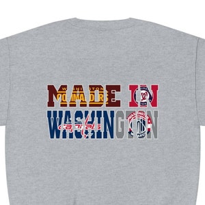 May include: A light gray sweatshirt with the text "MADE IN WASHINGTON" in a multi-colored design. The design incorporates logos and colors associated with Washington D.C. sports teams. The sweatshirt has long sleeves and a crew neck.