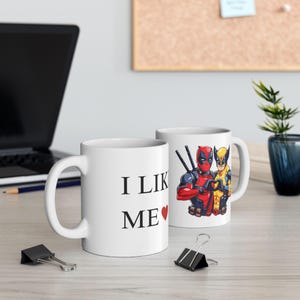 May include: Two white ceramic mugs with a graphic of Deadpool and Wolverine. One mug says "I Like Me" with a red heart.
