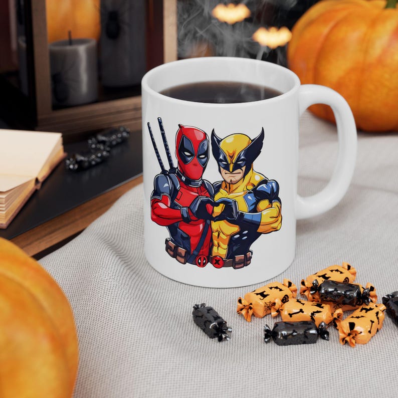 May include: White ceramic coffee mug with a graphic of Deadpool and Wolverine, both characters are wearing their signature costumes and are making a heart shape with their hands. The mug is on a white surface with a few candy pieces scattered around it.