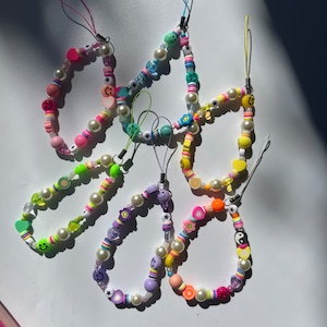 May include: A set of colorful beaded phone charms with various shapes and designs, including smiley faces, hearts, and stars. The charms are strung on white pearl beads and feature a variety of bright colors, including pink, yellow, green, blue, and purple.