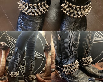 Op de afbeelding: Een paar zwarte leren cowboylaarzen met zilveren gesp-riemen. De laarzen hebben een spitse neus en een lage hak. De laarzen worden gedragen door een persoon met een zwarte broek.
