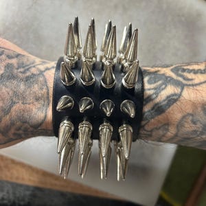 May include: Black leather wristband featuring rows of silver-colored metal spikes. The punk-inspired accessory has a bold, edgy design. The wristband is secured with metal snaps.