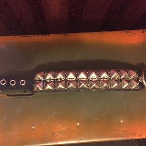 Puede incluir: Un brazalete de cuero negro con tachuelas de pirámide plateadas. El brazalete tiene un cierre de hebilla y múltiples ojales en la correa de cuero.
