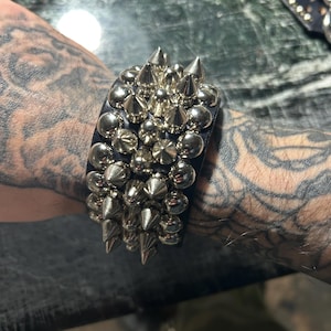 Puede incluir: Brazalete de cuero negro con púas y tachuelas de metal plateado. El brazalete está densamente cubierto de púas y tachuelas plateadas de varios tamaños, creando una estética audaz y vanguardista. Un accesorio llamativo.