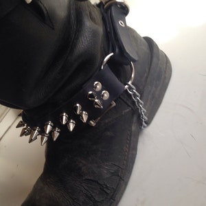 Könnte beinhalten: Schwarze Leder-Motorrad-Stiefel mit silbernen Nieten und einem Kettendetail. Die Stiefel haben einen abgenutzten Look und werden mit einer Schnalle geschlossen.