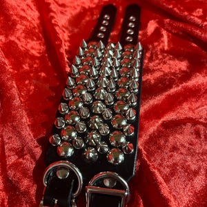 May include: Two black leather wristbands with rows of silver metal spikes and studs. Each wristband has a buckle closure and a D-ring. The wristbands are displayed on a red velvet surface.