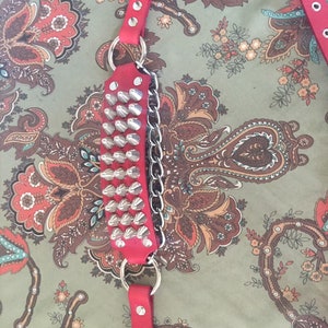 May include: Red leather studded belt with a silver chain and buckle. The belt is on a patterned fabric background.